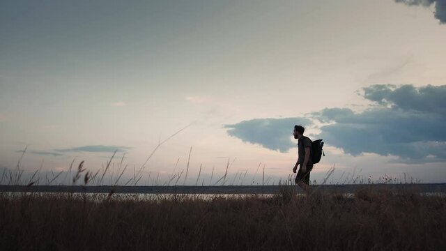 Man photographer with backpack is walking along meadow on plato of a hill and enjoying picturesque landscape and view on a river. Summer evening