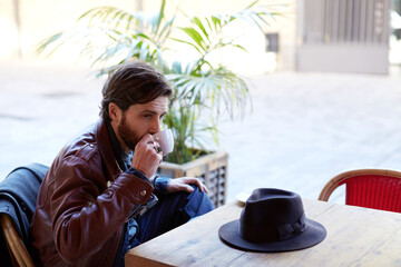 Portrait of handsome fashionable hipster man holding cup of tea or coffee next to his mouth while sitting on the terrace of beautiful coffee shop