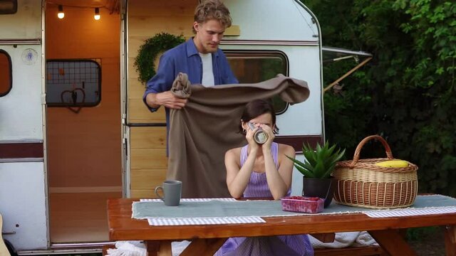 Young, european couple travelling, camping,living in trailer. Portrait of a woman dring a tea outdoors, while man coming to cover shoulder with plaid. Cool morning, outdoors