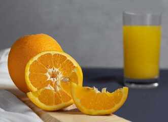 An orange half, an orange and its slices on a wooden stand, a glass of orange juice out of focus, on a gray background.