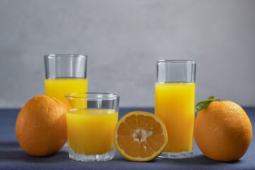 Homemade orange juice in different glasses with oranges nearby on a gray background.