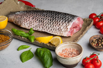 Headless carp carcass, peeled from fish scales, with lemon, cherry tomatoes, pepper and spices on a large clay dish on a gray background