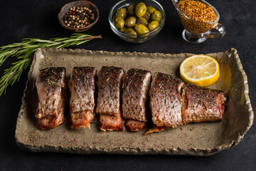 Baked carp with greens, lemon, olives and spices on a clay plate on a black background