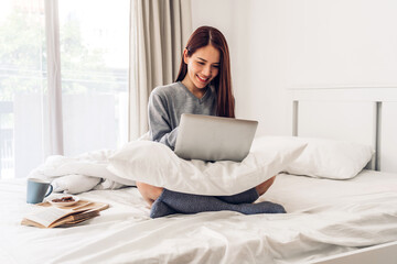 Obraz premium Young smiling happy beautiful asian woman relaxing using laptop computer in the bedroom at home. Young creative girl working and typing on keyboard.work from home concept
