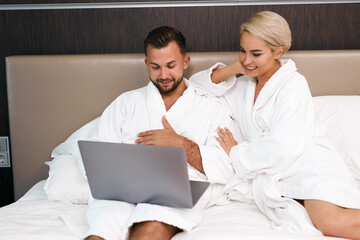 Obraz premium young caucasian couple with laptop in the morning, bearded man and blonde woman discuss where to go during their trip, they lie on bed in bathrobes, in hotel