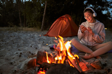 Cheerful young woman using cellphone while sitting neat campfire