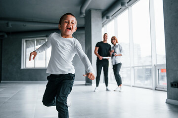 Fototapeta premium Woman in formal clothes and with camera in hand standing inside of empty room with man and little boy that running and having fun