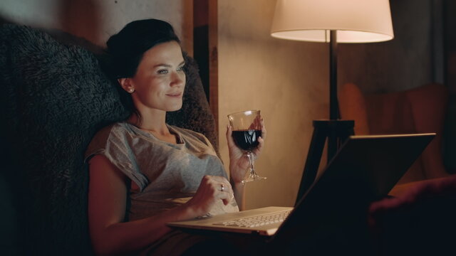 Woman drinking red wine with notebook. Girl looking laptop with wineglass.