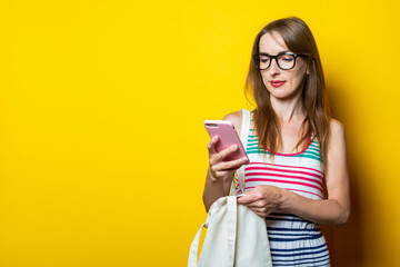 Beautiful young girl in glasses looks at the phone, with a linen bag on her shoulder on a yellow background.