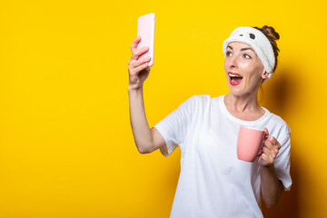 Young woman in a bandage for sleeping and with a cup of coffee makes a selfie, posing on a yellow background