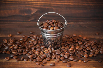 Coffee beans in small metal bucket on wooden surface. Beans scattered around. Close-up.