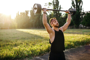 Male athlete lifting a barbell overhead, training an athlete outdoors at sunset, sports equipment...