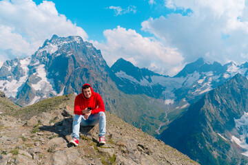 Fototapeta premium Man tourist sitting on rock with smartphone in mountains. Male traveler using mobile phone against cloudy sky on sunny day in mountainous terrain.