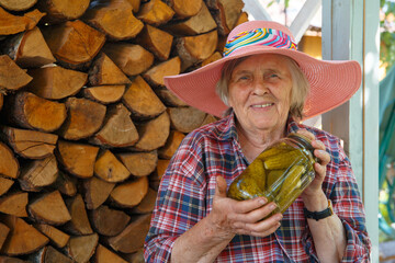 Fototapeta premium An elderly woman in a pink hat is holding a jar of cucumbers. Hand-made canned cucumbers.