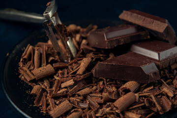 Close-up pieces of chocolate, chocolate shavings and peeler in black plate on dark textured background.