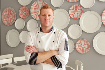 Young handsome tall redhead blue-eyed men chef cook stands with his arms crossed against the background of the wall of his restaurant decorated with plates