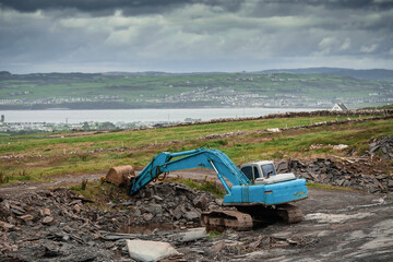 Fototapeta na wymiar Construction site in a field with blue color excavator working. Dark dramatic sky, West of Ireland.