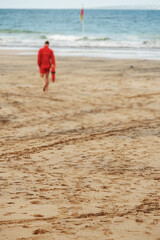 Fototapeta na wymiar Ocean sand in focus with many foot prints. Lifeguard walking towards a flag in water out of focus Concept end of busy day at a beach.