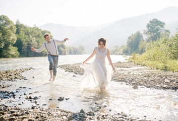 Fototapeta na wymiar happy, emotional gorgeous bride and stylish groom having fun, splashed with water. Wedding couple running along the mountain river with bare feet.