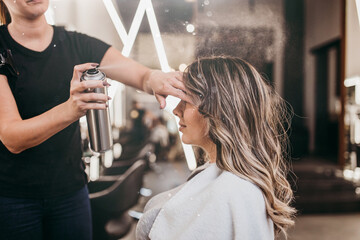 Beautiful young woman with long curly hair in hair salon. Professional hairdresser styling with hairspray. - Powered by Adobe