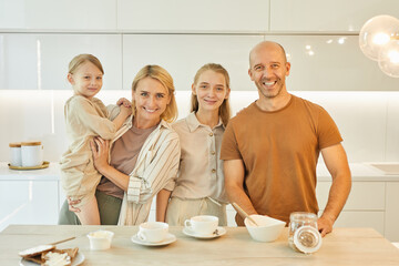 Warm-toned waist up portrait of modern happy family looking at camera while enjoying breakfast together standing by table in minimal kitchen interior, copy space