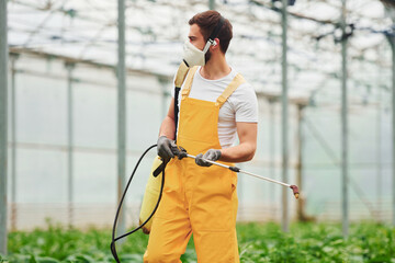 Young greenhouse worker in yellow uniform and white protective mask watering plants by using special equipment inside of hothouse