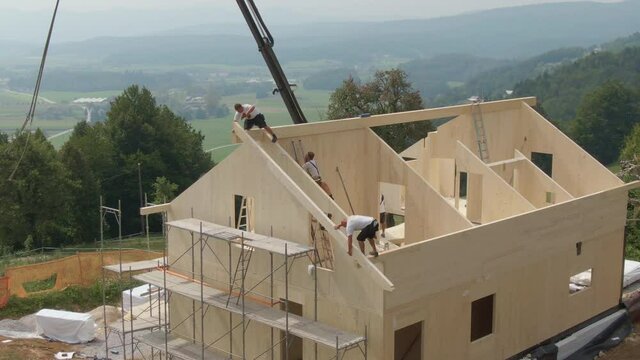 DRONE: Flying around a modern CLT house being built in the country by courageous builders. Construction site workers walks along a wooden beam running across the cross-laminated timber house's roof.