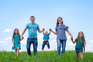 Big family dad, mom and three children walk on green grass against blue sky. Happy caucasian parents, two daughters and young son holding hands.
