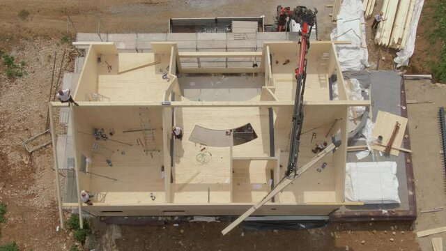 TOP DOWN: Flying above a modern cross-laminated timber house under construction as a large boom lifts a long wooden beam up to the contractors working on the roof. Building of a prefabricated house.