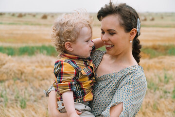 Fototapeta na wymiar Beautiful young woman with fair-haired little son in her arms in the middle of a wheat field with bales in summer evening