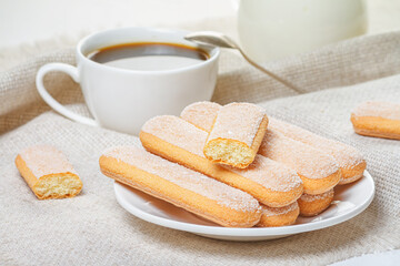 Traditional savoiardi biscuits or ladyfingers cookies on a white plate and a white cup of coffee with a spoon on a napkin. Selective focus
