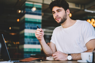Talented young student hipster guy dressed in casual white t shirt looking out of window holding...
