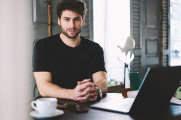 Portrait of cheerful young man working freelance in cafeteria interior while looking at camera with smile on face.Positive handsome student enjoying leisure time sitting with laptop computer indoors