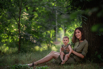 A healthy young mother holds a toddler baby in her arms. A happy family sits on the green grass, under a tall tree, plays, hugs, enjoys a walk in the park. The concept of motherhood, childcare.
