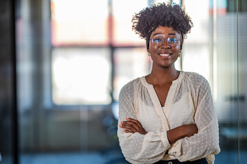 Pretty young dreamy African-American office worker standing with arms crossed and looking at camera. Young business woman in modern office. Young African ethnicity businesswoman