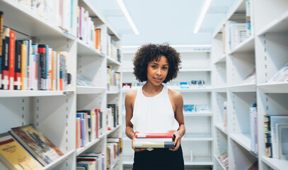 Portrait of attractive dark skinned woman student spending free time in library holding favorite fiction books, beautiful curly african american female looking at camera standing near shelves in store - obrazy, fototapety, plakaty