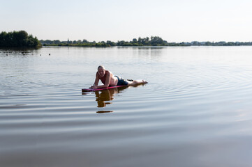 Elderly man lying on a surfboard in a lake with reflective water in the evening twilight.