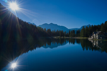 Sun on a crystal blue sky. Reflection of the sun in the water. Early morning in the Alps. House in the mountains by a mountain lake. Reflections of trees, mountains, sky in a mountain lake.
