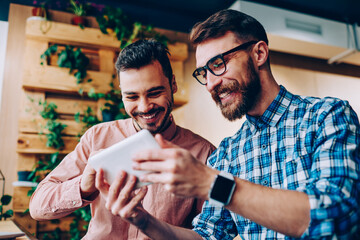Cheerful young hipster guys having fun watching funny videos from social networks via touchpad together, smiling bearded man showing his results to friend playing online games on portable pc