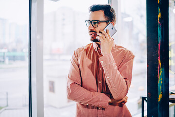 Pensive young man in casual wear looking away waiting for connection with service operator calling via smartphone,dissatisfied guy having telephone conversation about problem standing indoors.