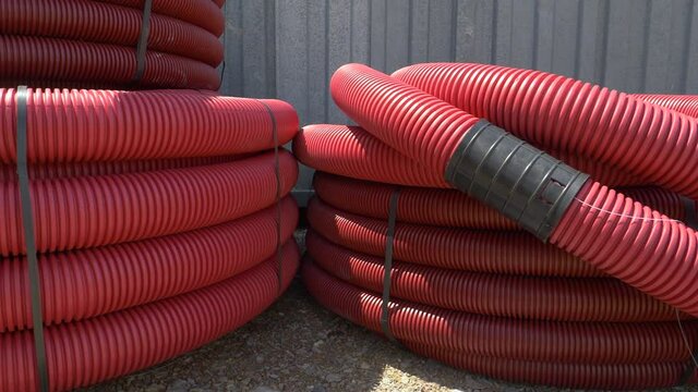 CLOSE UP: Hot summer sunbeams shine on two stacks of pvc sewage tubing placed near a house under construction. Two large coils of red plastic conduits lay near a busy construction site. Utility tubes