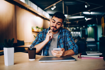 Contemplative male analyzing information about booking on website using cellular in coworking...