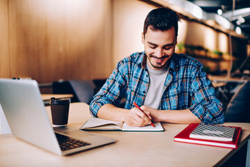 Smiling male blogger satisfied with having great idea for publication making notes sitting at desktop with netbook, positive student enjoying learning process doing homework task in college library.