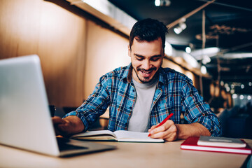 Positive smiling male student writing essay with excited idea in textbook for education while sitting with modern laptop computer in college campus and enjoying leisure time for autodidact indoors