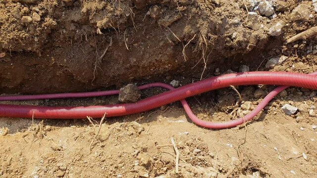 CLOSE UP: Wide red plastic telecommunication conduits run along a newly dug up shallow trench. Corrugated plastic utility tubing lies inside a trench dug into the soil of a road under construction.
