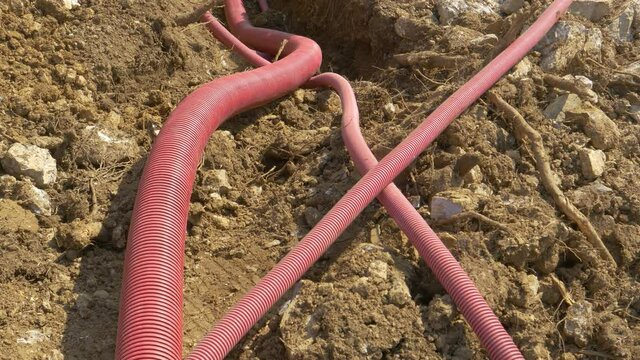 CLOSE UP: Corrugated plastic utility tubing lies inside a trench dug into the soil of a road under construction. Wide red plastic telecommunication conduits run along a newly dug up shallow ditch.