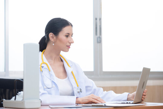 Female doctor with stethoscope sitting at table use computer laptop  in office hospital. Confident woman doctor sitting at office desk, health care and prevention technology concept