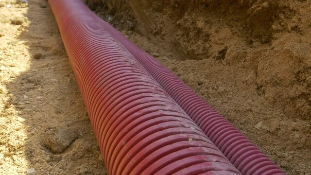 CLOSE UP: Detailed shot of corrugated plastic telecommunication tubing in a deep ditch running next to a road under construction. Ridged waterproof conduit runs along a trench dug in light brown soil.