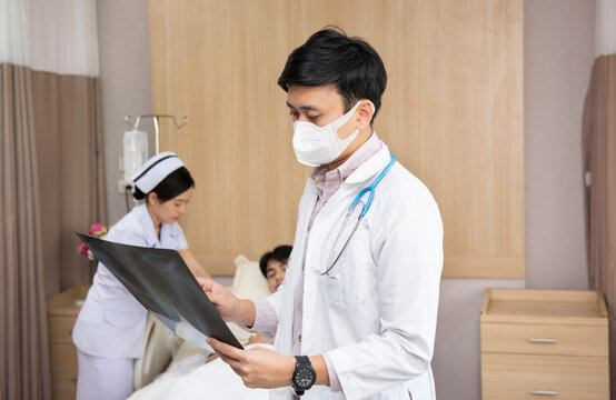 Asian Doctor With Face Mask Checking Lab Test Film X-ray Results Of Elderly Hospice Patient With Nurse At Hospital. People Health And Medical Concept.