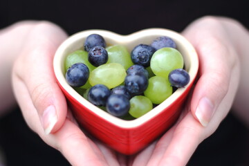 hands holding heart shape dish with tasty fresh grapes and blueberries